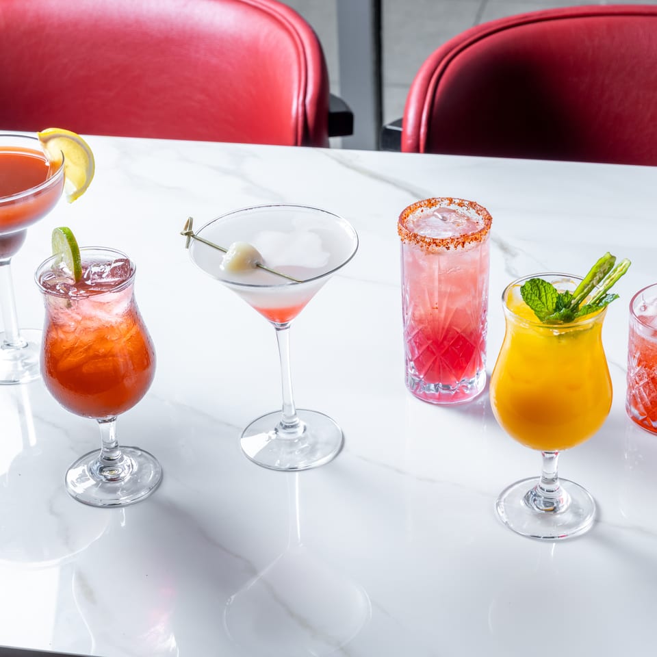 Colorful cocktails in assorted glasses sit on a white marble restaurant table with red upholstered chairs.