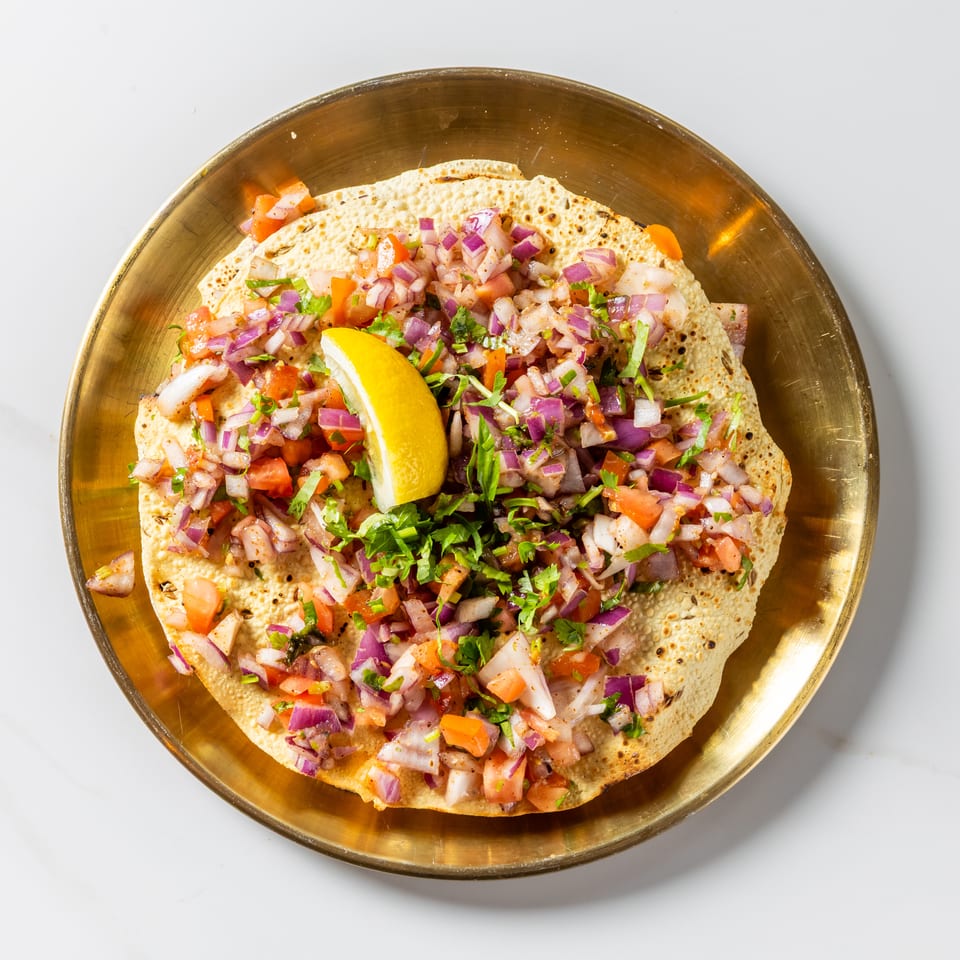 Crisp flatbread topped with diced tomatoes, red onions, herbs, and a lemon wedge on a brass plate.