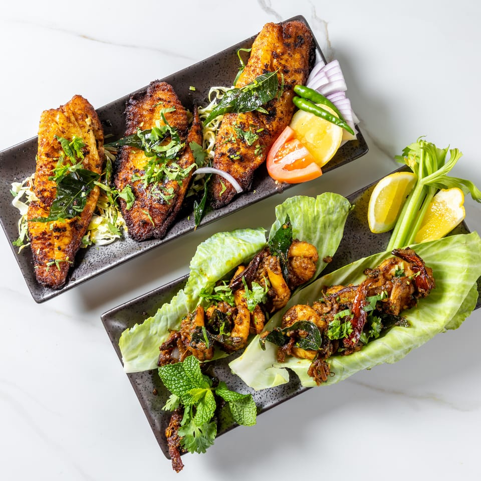 Grilled spiced fish and seafood served on black platters with lettuce, herbs, lemon, tomato, onion, and chilies.