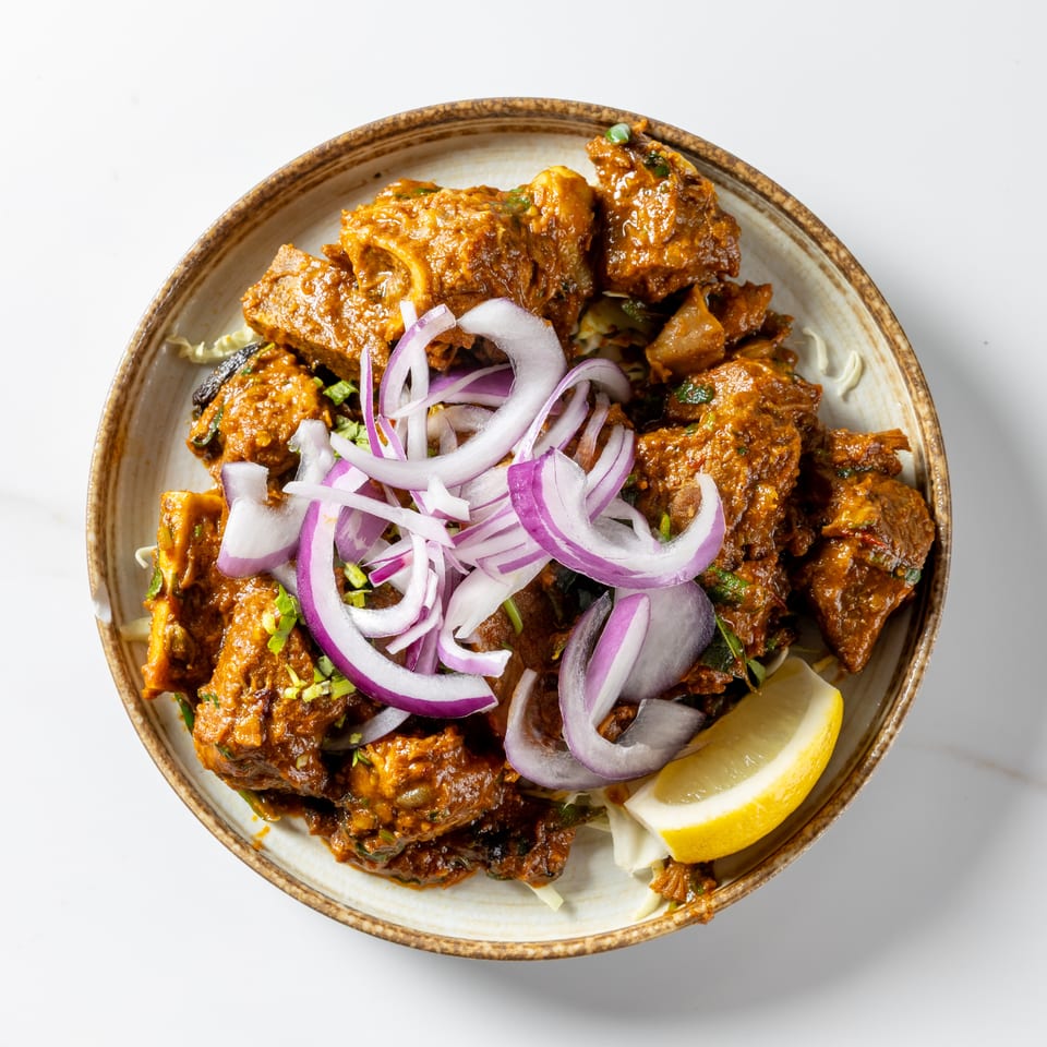Overhead view of spiced meat curry garnished with sliced red onions, herbs, cabbage, and a lemon wedge.