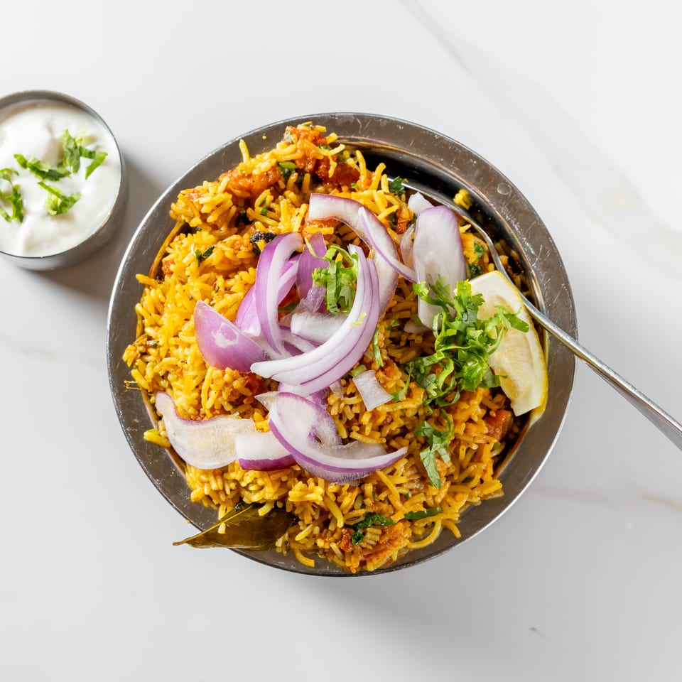 Overhead view of spiced biryani rice topped with red onions, cilantro, and lemon, served with yogurt sauce.