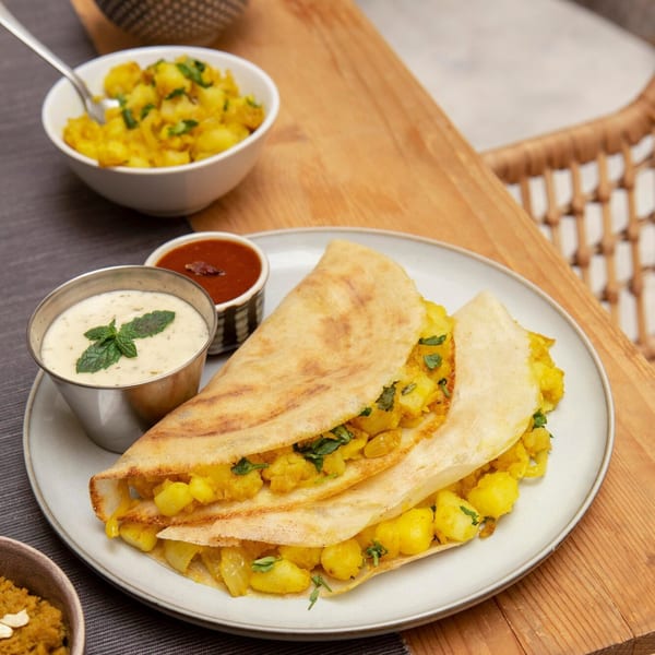 Two golden-brown dosas filled with spiced potatoes, served with chutney and raita on a wooden table.