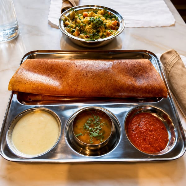 A golden-brown dosa with three dipping sauces on a metal tray, with a vegetable dish in the background.