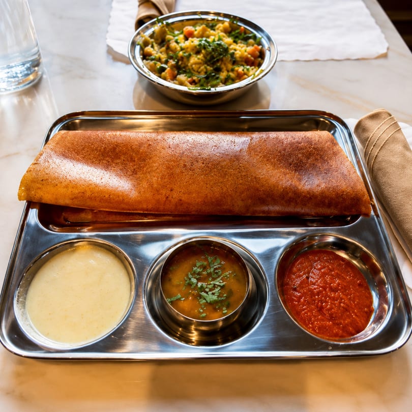 A golden-brown dosa with three dipping sauces on a metal tray, with a vegetable dish in the background.