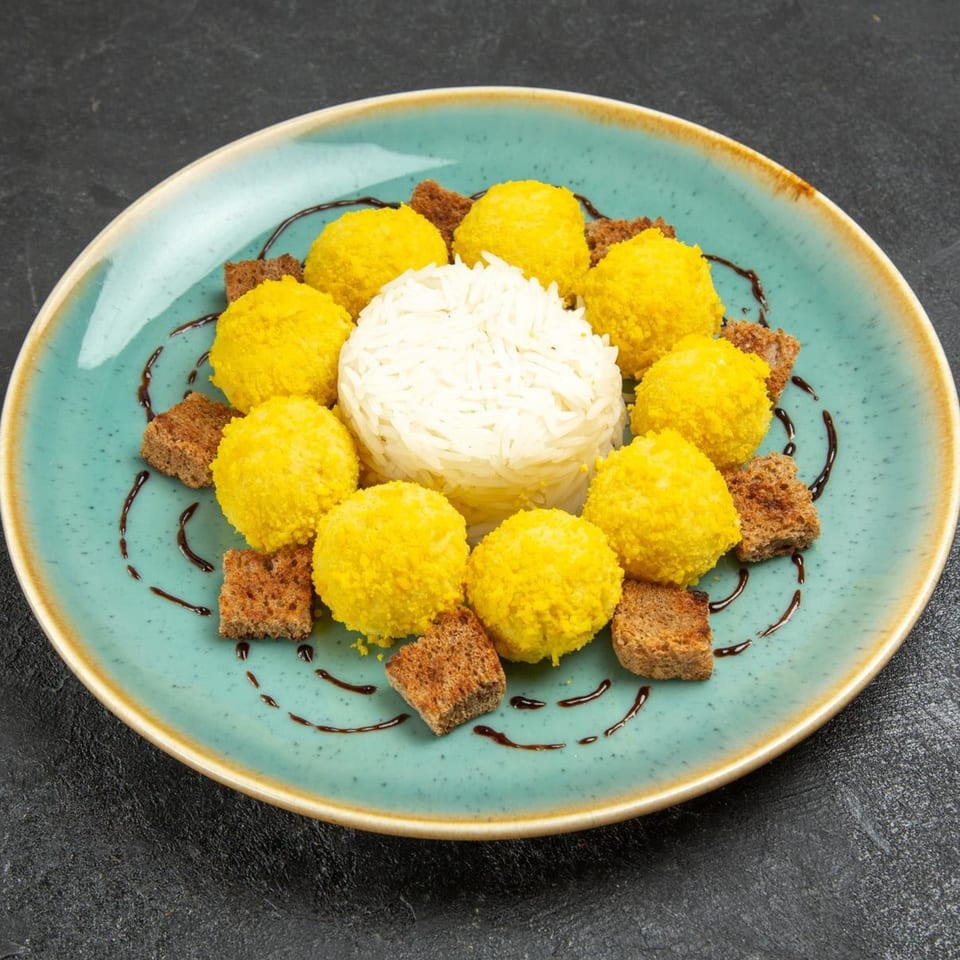 A teal plate with a mound of white rice surrounded by yellow fritters and brown croutons, drizzled with sauce.