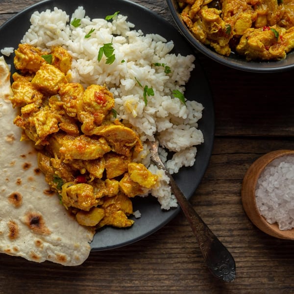Indian chicken curry with rice and naan bread on a black plate, with a bowl of salt.