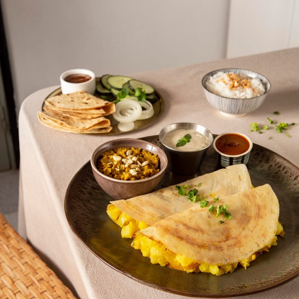 Indian dosa with potato filling, chutneys, rice, flatbreads, and sliced vegetables on a table.
