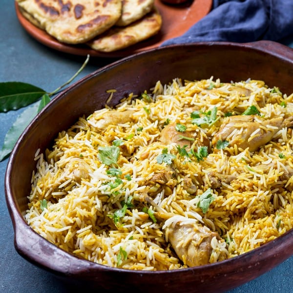 Chicken biryani with cilantro in a rustic bowl, with naan bread and bay leaves in the background.